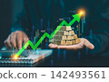 Businessman holding stack of gold bullion bars with green rising stock graph and candlestick chart overlay, gold investment and wealth concept. 142493561