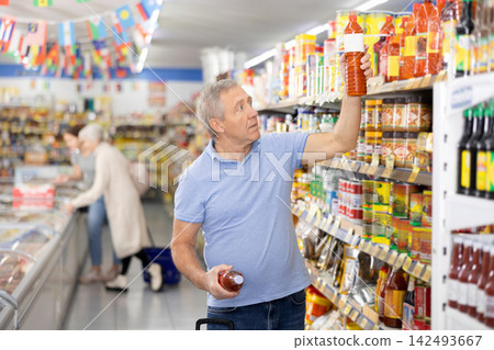 Elderly man chooses and buys hot chili sauce in the grocery section of supermarket Elderly man chooses and buys hot chili sauce in the grocery section of supermarket 142493667