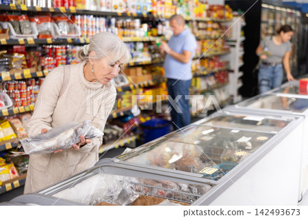 Old woman purchaser choosing frozen fish in big supermarket Old woman purchaser choosing frozen fish in big supermarket 142493673