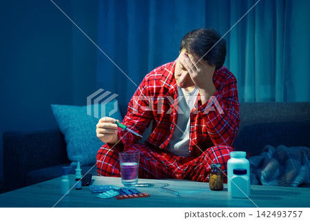 man with flu symptoms checks his fever with a thermometer. He uses cold medicine for home treatment, focusing on recovery from a viral infection and illness. 142493757