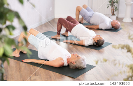 Class of elderly sporty women stretching gluteal muscle while lying on back during yoga Pilates workout on mats in gym studio Class of elderly sporty women stretching gluteal muscle while lying on back during yoga Pilates workout on mats in gym studio 142493993