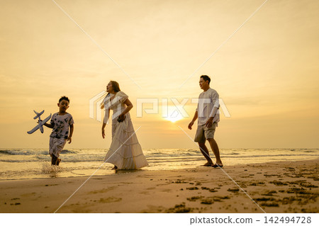 Family walking together on beach during sunset with child holding toy airplane, enjoying tropical vacation outdoors near ocean waves during summer evening trip for joy Family walking together on beach during sunset with child holding toy airplane, enjoying tropical vacation outdoors near ocean waves during summer evening trip for joy 142494728