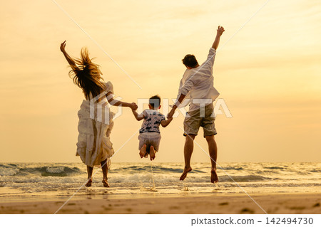 Family jumping together on beach during sunset while enjoying fun holiday vacation outdoors with joyful people holding hands near ocean water with warm sunlight lighting up scenery Family jumping together on beach during sunset while enjoying fun holiday vacation outdoors with joyful people holding hands near ocean water with warm sunlight lighting up scenery 142494730