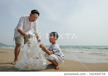 Family picking up plastic garbage in transparent bag during holiday vacation on sandy beach, father and son helping ecology by cleaning rubbish near ocean waves outdoors Family picking up plastic garbage in transparent bag during holiday vacation on sandy beach, father and son helping ecology by cleaning rubbish near ocean waves outdoors 142494739