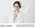 一位中年婦女雙手抱頭，在白色背景下看著她的智慧型手機。 142495038