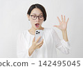 一位中年婦女在白色背景下驚訝地看著她的智慧型手機。 142495045