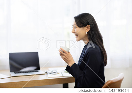 A young Japanese businesswoman in a suit sits at a desk by a bright window, coffee cup in hand, talking to a colleague. A young Japanese businesswoman in a suit sits at a desk by a bright window, coffee cup in hand, talking to a colleague. 142495654