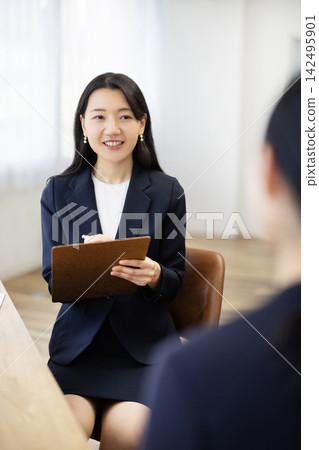 A young Japanese businesswoman in a suit, smiling and taking notes at a desk by a bright window, talks to a client. A young Japanese businesswoman in a suit, smiling and taking notes at a desk by a bright window, talks to a client. 142495901