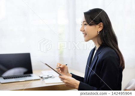一位身著職業套裝的年輕日本女商人，面帶微笑，坐在明亮的窗邊辦公桌前做筆記，正在與客戶交談。 142495904