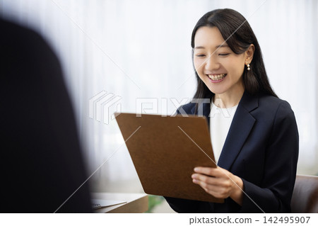 一位身著職業套裝的年輕日本女商人,面帶微笑,坐在明亮的窗邊辦公桌前做筆記,正在與客戶交談。 一位身著職業套裝的年輕日本女商人,面帶微笑,坐在明亮的窗邊辦公桌前做筆記,正在與客戶交談。 142495907