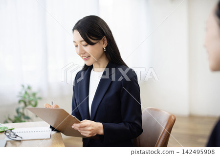 A young Japanese businesswoman in a suit, smiling and taking notes at a desk by a bright window, talks to a client. A young Japanese businesswoman in a suit, smiling and taking notes at a desk by a bright window, talks to a client. 142495908
