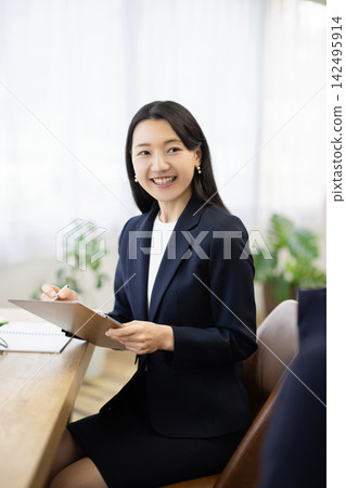 一位身著職業套裝的年輕日本女商人,面帶微笑,坐在明亮的窗邊辦公桌前做筆記,正在與客戶交談。 一位身著職業套裝的年輕日本女商人,面帶微笑,坐在明亮的窗邊辦公桌前做筆記,正在與客戶交談。 142495914