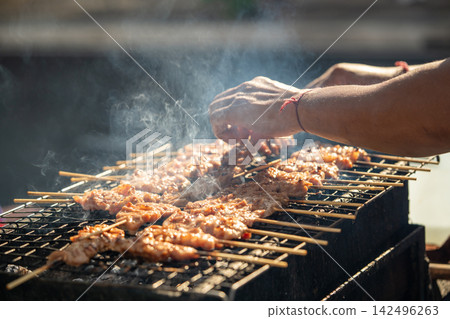 Thai man grilling chicken skewers over smoky charcoal barbecue at an outdoor street food stall. Thai man grilling chicken skewers over smoky charcoal barbecue at an outdoor street food stall. 142496263