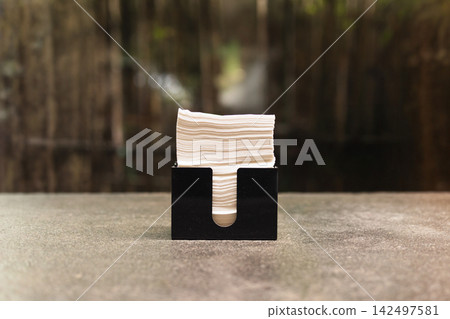 Stack of white tissue paper napkins inside a sleek black holder placed on a stone table in a cafe. Stack of white tissue paper napkins inside a sleek black holder placed on a stone table in a cafe. 142497581