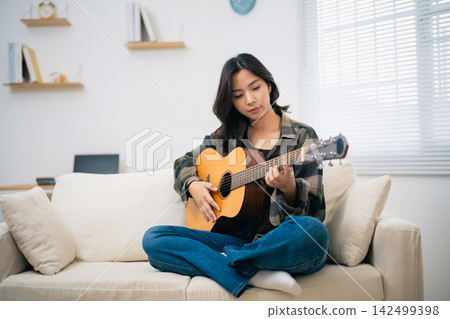 Young woman sitting on beige sofa playing acoustic guitar in cozy modern living room with natural light 142499398