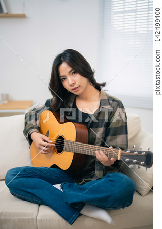 Young woman sitting cross-legged on sofa playing acoustic guitar with focused expression at home in casual outfit Young woman sitting cross-legged on sofa playing acoustic guitar with focused expression at home in casual outfit 142499400