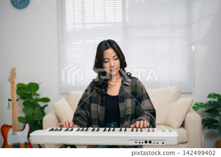 Young woman playing electronic keyboard indoors with focused expression sitting in cozy room with plants and guitar in background 142499402