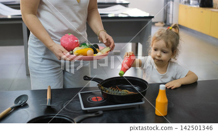 Little girl playing together with mother at a play kitchen preparing a dish with toy seafood and vegetables. Family cooking play concept Little girl playing together with mother at a play kitchen preparing a dish with toy seafood and vegetables. Family cooking play concept 142501346