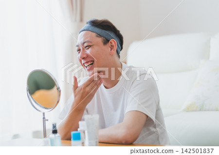 A 40-year-old man smiles in front of the mirror while touching his chin after skincare. (Men's beauty) 142501837