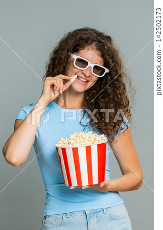 Woman watching exciting movie in 3D glasses eating popcorn, emotional surprised amazed reaction 142502173