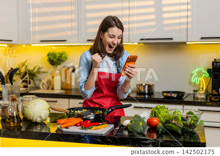 Young woman celebrating prize win on smartphone indoors in modern home kitchen while healthy cooking Young woman celebrating prize win on smartphone indoors in modern home kitchen while healthy cooking 142502175