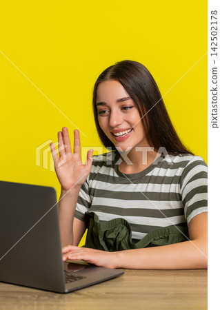 Woman smiling and waving hand to webcam on laptop during online chat at table joyful connection Woman smiling and waving hand to webcam on laptop during online chat at table joyful connection 142502178