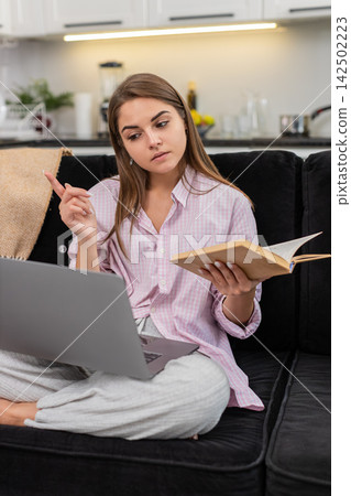 Young woman watches online course on laptop with book nearby nods to teacher explanation and learns Young woman watches online course on laptop with book nearby nods to teacher explanation and learns 142502223