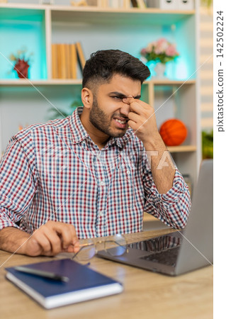 Indian man on home table rubbing tired eyes from laptop work and feeling fatigue during remote job Indian man on home table rubbing tired eyes from laptop work and feeling fatigue during remote job 142502224