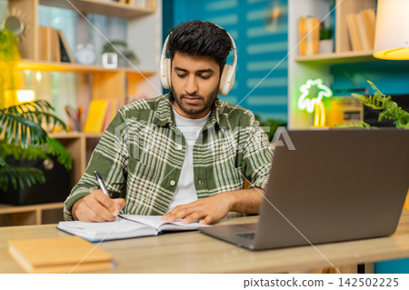 Indian man on home table taking notes in headphones during online lesson with serious focused face Indian man on home table taking notes in headphones during online lesson with serious focused face 142502225