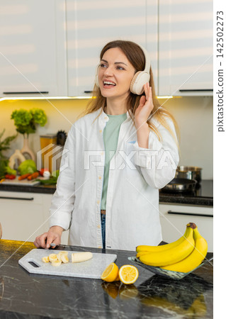 Middle-aged woman listening to music while slicing banana for dietary smoothie snack in home kitchen 142502273