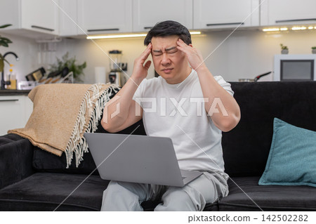 Asian man with laptop feels headache rubs temples closes eyes breathes slowly takes short work break Asian man with laptop feels headache rubs temples closes eyes breathes slowly takes short work break 142502282