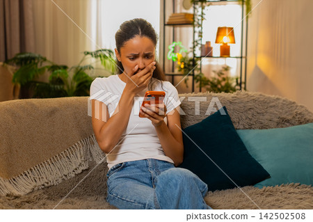 African American young woman on home sofa sighing with disappointment after bad news on smartphone African American young woman on home sofa sighing with disappointment after bad news on smartphone 142502508