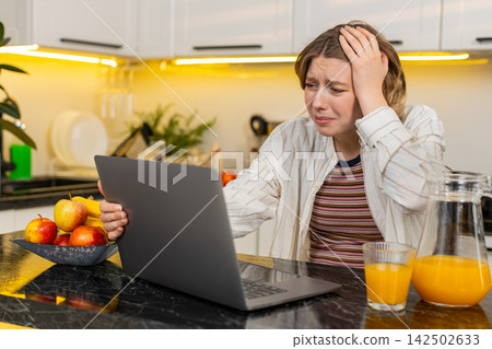 Young woman feeling stressed by laptop in home kitchen during diet break after upsetting online news 142502633