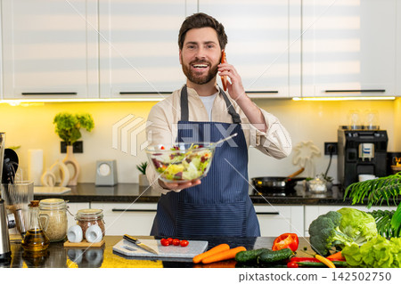 Middle-aged man tasting fresh vegetable salad while talking on smartphone in home kitchen for diet Middle-aged man tasting fresh vegetable salad while talking on smartphone in home kitchen for diet 142502750