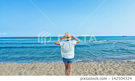 A woman wearing a straw hat, seen from behind, gazing at the sea. A woman wearing a straw hat, seen from behind, gazing at the sea. 142504324