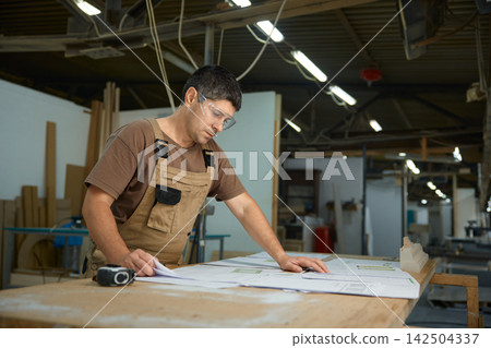 Workman assessing construction drawings in workshop while using measuring instruments and tools Workman assessing construction drawings in workshop while using measuring instruments and tools 142504337