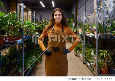 Indoor plant shop gardener holding trowel, apron and gloves surrounded by potted foliage 142504367