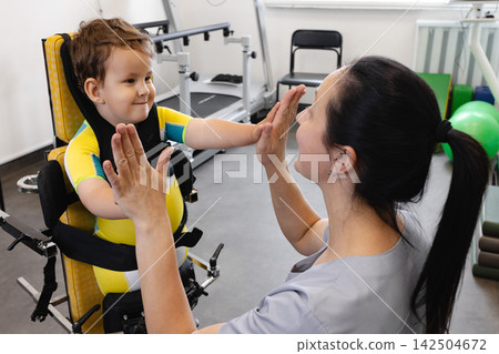 Happy boy during physical rehabilitation standing in a standing frame with his physical therapist, working on developing standing skills Happy boy during physical rehabilitation standing in a standing frame with his physical therapist, working on developing standing skills 142504672