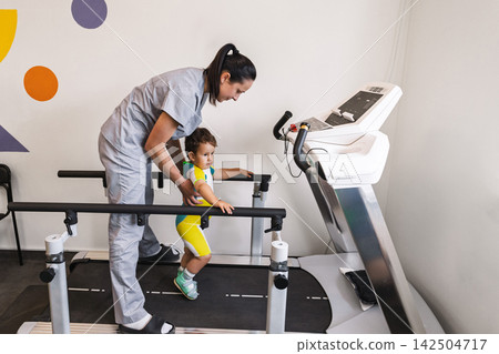 Child wearing rehab clothes and shoes is receiving help while using a treadmill in a rehabilitation clinic for therapy Child wearing rehab clothes and shoes is receiving help while using a treadmill in a rehabilitation clinic for therapy 142504717