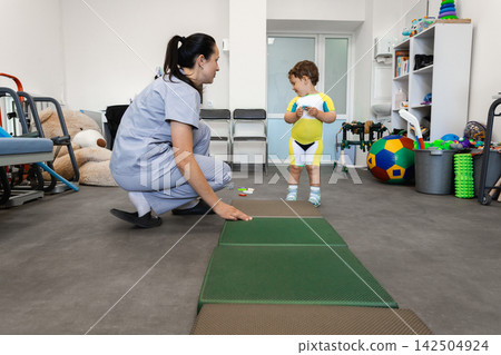 Child after an injury in a rehabilitation center where woman physical therapist uses the balance-pad solid for rehabilitation and balance and coordination training Child after an injury in a rehabilitation center where woman physical therapist uses the balance-pad solid for rehabilitation and balance and coordination training 142504924