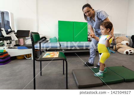 Children's rehabilitation. Boy with a disability walks on balance pads under the supervision of a physical therapist, to train and improve walking and balance skills Children's rehabilitation. Boy with a disability walks on balance pads under the supervision of a physical therapist, to train and improve walking and balance skills 142504927
