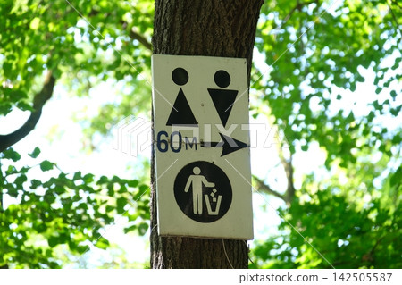 Public Toilet and Trash Bin Direction Sign Mounted on Tree Showing Restroom Symbols Arrow and Distance in Green Outdoor Park Public Toilet and Trash Bin Direction Sign Mounted on Tree Showing Restroom Symbols Arrow and Distance in Green Outdoor Park 142505587