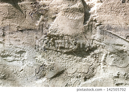 Rough beige concrete wall with shallow cracks, worn patches and uneven relief shows natural aging and weathering of a construction material. Rough beige concrete wall with shallow cracks, worn patches and uneven relief shows natural aging and weathering of a construction material. 142505702