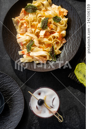 Noodle plate. Cocktail drink. Tasty spaghetti with salmon red caviar in cream sauce. Delicious restaurant dining set on black table. 142506359