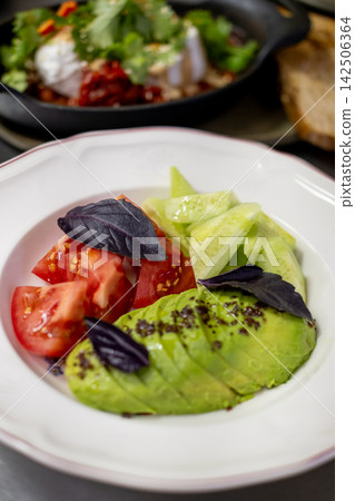 Salad plate. Healthy vegetable breakfast. Sliced avocado with spices red tomato cucumber on white plate served with purple basil leaves. Salad plate. Healthy vegetable breakfast. Sliced avocado with spices red tomato cucumber on white plate served with purple basil leaves. 142506364