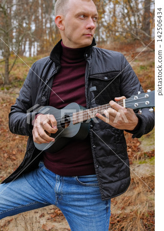 Man plays ukulele while spending time outdoors in autumnal natural setting. Musician holds small string instrument and performs music in relaxed atmosphere surrounded by greenery and open space Man plays ukulele while spending time outdoors in autumnal natural setting. Musician holds small string instrument and performs music in relaxed atmosphere surrounded by greenery and open space 142506434