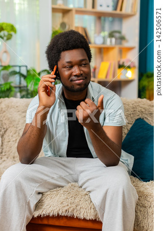 African American young man on home sofa answering smartphone call during calm talk at home interior African American young man on home sofa answering smartphone call during calm talk at home interior 142506571