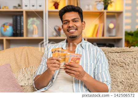 Indian man on home sofa counting cash money in living room with satisfied smile and personal success Indian man on home sofa counting cash money in living room with satisfied smile and personal success 142506572