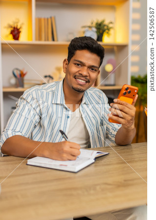 Indian man on home table holding smartphone and writing notes in notebook during focused remote work Indian man on home table holding smartphone and writing notes in notebook during focused remote work 142506587