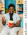 African American young man on home sofa watching sports with popcorn and cheering for match win joy 142506599